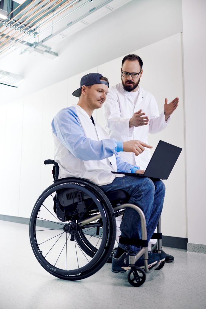 Ein Mann mit Labor-Kittel im Rollstuhl spricht mit einem anderen Mann in einem Labor.